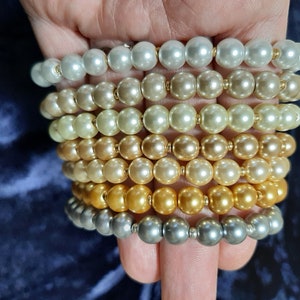 Op de afbeelding: Een stapel parel armbanden in verschillende tinten wit, crème, goud en zilver. De armbanden zijn gemaakt van nepparels en zijn gerangschikt in een aflopende volgorde van grootte, met de grootste parels onderaan en de kleinste bovenaan.