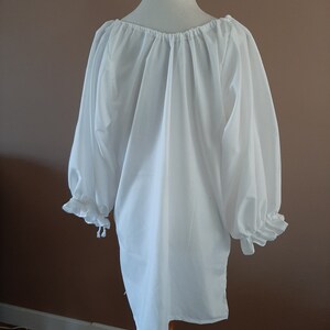 May include: White peasant blouse with long sleeves and a gathered neckline. The sleeves have ruffles at the cuffs and are tied at the wrists.
