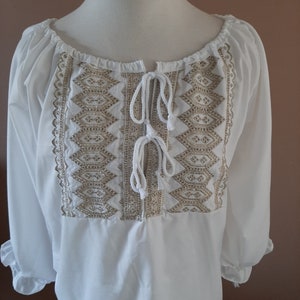 May include: White peasant blouse with a gold embroidered design and a tie at the neckline.