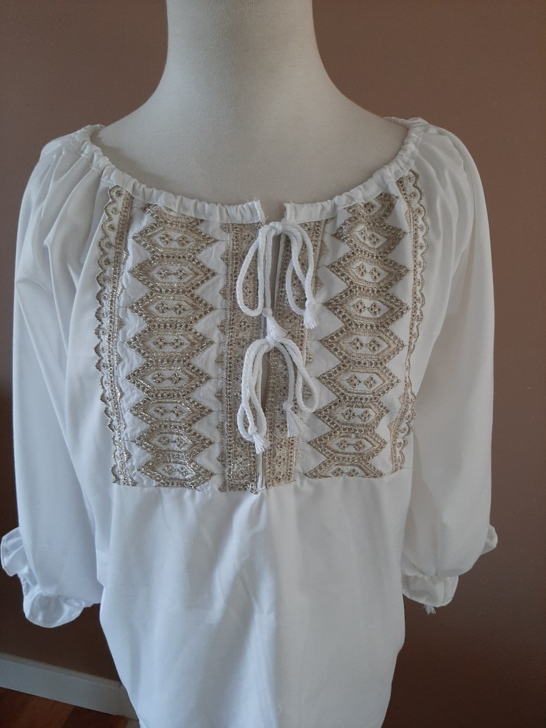 May include: White peasant blouse with a gold embroidered design and a tie at the neckline.