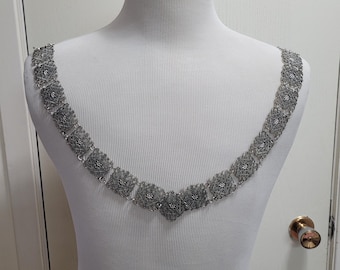 May include: A silver-toned chain necklace featuring a series of square, intricate filigree links connected by small circular jump rings. The necklace is draped in a V-shape, showcasing a repeating pattern of detailed metalwork across each square segment.