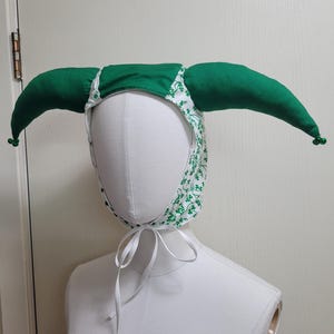 Pode incluir: Um chapéu de bobo da corte com dois chifres longos de tecido verde acolchoado que se estendem pelas laterais, cada um com um pequeno sino verde na ponta. A base é de tecido branco com estampa de trevos verdes e fitas brancas para amarrar.