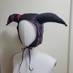 Pode incluir: Um gorro estilo bobo da corte com dois chifres longos e acolchoados nas laterais. O tecido apresenta um padrão preto, vermelho e azul com um motivo de losangos. O gorro inclui laços finos roxo-escuros e pequenas contas nas extremidades.