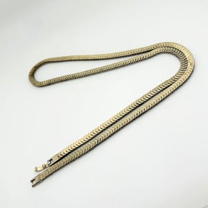 May include: A gold-toned herringbone chain necklace featuring a flat, flexible design with a repeating chevron pattern. The necklace includes a rectangular box clasp closure at the end, showcasing a polished metallic finish throughout the entire piece.
