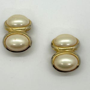 May include: A pair of vintage clip-on earrings. Each earring features a faux pearl set in a gold-tone and silver-tone metal setting. The design is a stacked oval shape.