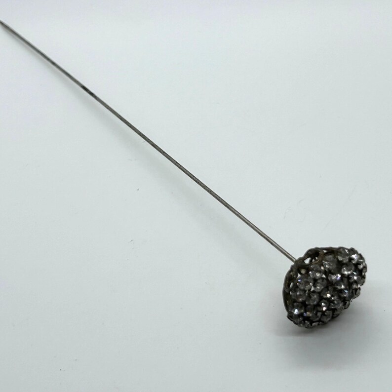 May include: A vintage hat pin with a silver-toned metal shaft and a cluster of sparkling, clear rhinestones forming the head. The pin is designed to secure hats or other accessories.