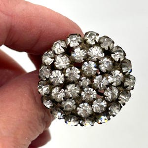 May include: A round, vintage brooch with a cluster of clear, sparkling rhinestones. The rhinestones are set in a tarnished, possibly antique, metal base. The brooch is held by a hand, suggesting its size and wear.