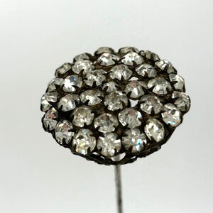 May include: A vintage hat pin with a cluster of clear, faceted rhinestones. The rhinestones are set in a dark metal base, creating a sparkling, dome-shaped design. The pin has a long, slender silver-toned stem.