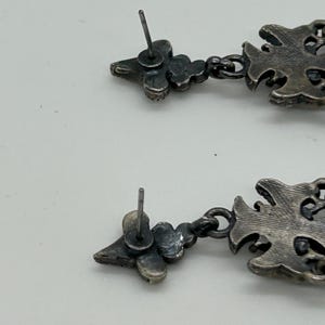 Puede incluir: Un par de pendientes plateados. Cada pendiente presenta un perno con un dise&ntilde;o floral, conectado a un colgante calado decorativo. Los colgantes tienen un patr&oacute;n ornamental y sim&eacute;trico. Los pendientes est&aacute;n dise&ntilde;ados para orejas perforadas.