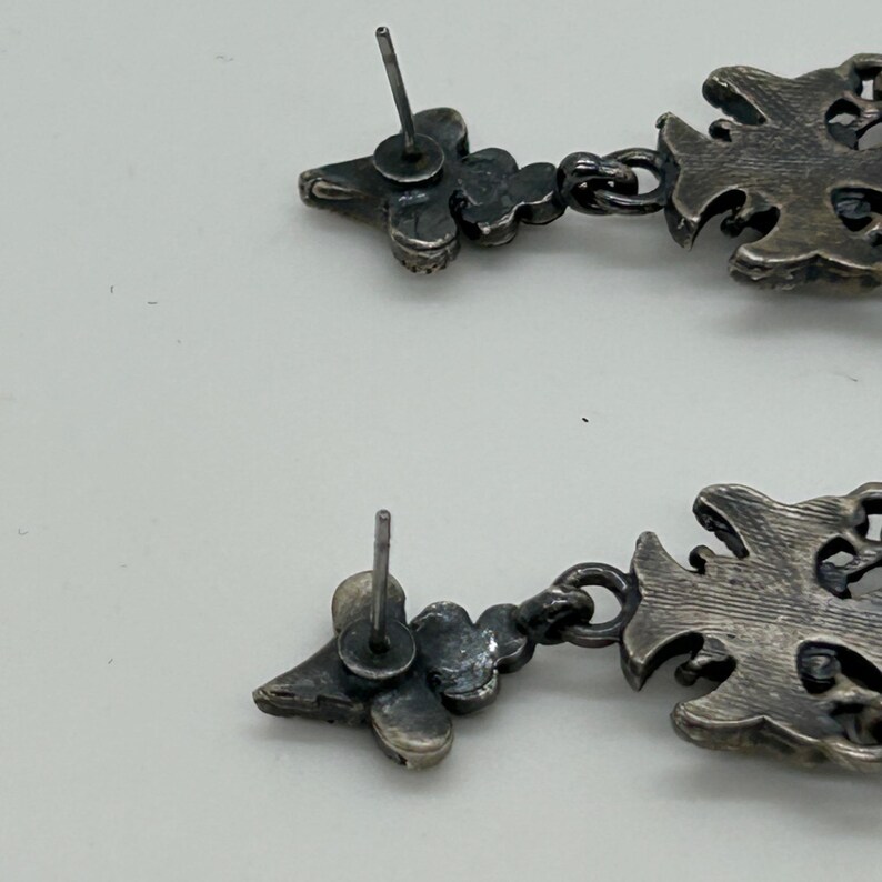Puede incluir: Un par de pendientes plateados. Cada pendiente presenta un perno con un dise&ntilde;o floral, conectado a un colgante calado decorativo. Los colgantes tienen un patr&oacute;n ornamental y sim&eacute;trico. Los pendientes est&aacute;n dise&ntilde;ados para orejas perforadas.