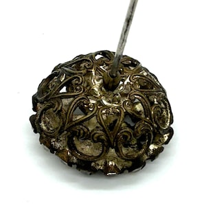 May include: Close-up of an antique silver-toned hat pin. The pin features an ornate, openwork design with swirling patterns and a central mounting for the pin itself. The metal has a tarnished appearance, suggesting age.
