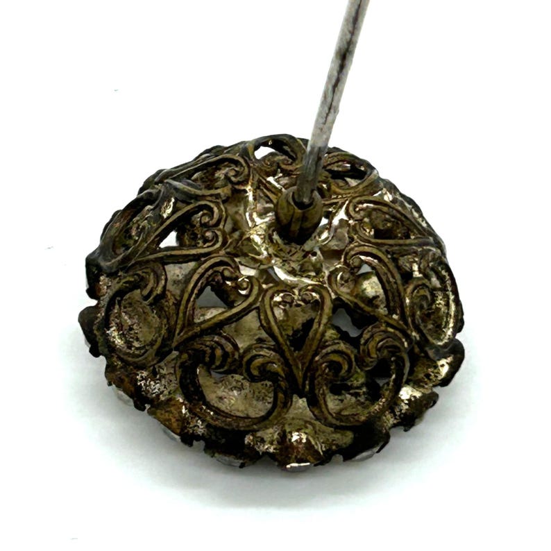 May include: Close-up of an antique silver-toned hat pin. The pin features an ornate, openwork design with swirling patterns and a central mounting for the pin itself. The metal has a tarnished appearance, suggesting age.