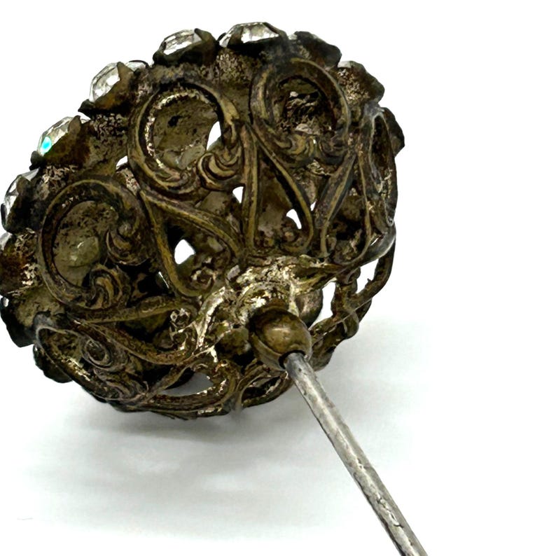 May include: Antique silver-toned hat pin with a filigree design. The pin's head is adorned with clear, faceted crystals around the edge. The intricate metalwork and sparkling crystals create a vintage aesthetic.