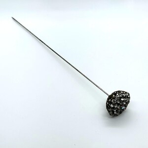 May include: A silver-toned hat pin with a decorative head. The head is oval-shaped and encrusted with small, sparkling crystals. The pin is long and slender, designed to secure a hat or other headwear.