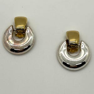 May include: A pair of clip-on earrings with a gold-colored top and a silver-colored, half-circle base. The earrings have a vintage look and are set against a white background.