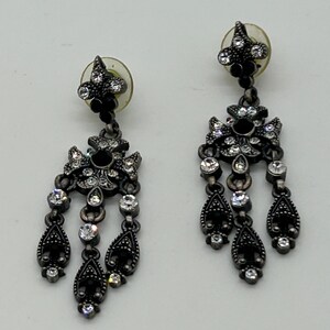 Puede incluir: Par de pendientes colgantes grises oscuros y ornamentados. Cada pendiente presenta un dise&ntilde;o floral con detalles de cristal negro y transparente. Los pendientes tienen un dise&ntilde;o en forma de l&aacute;grima.