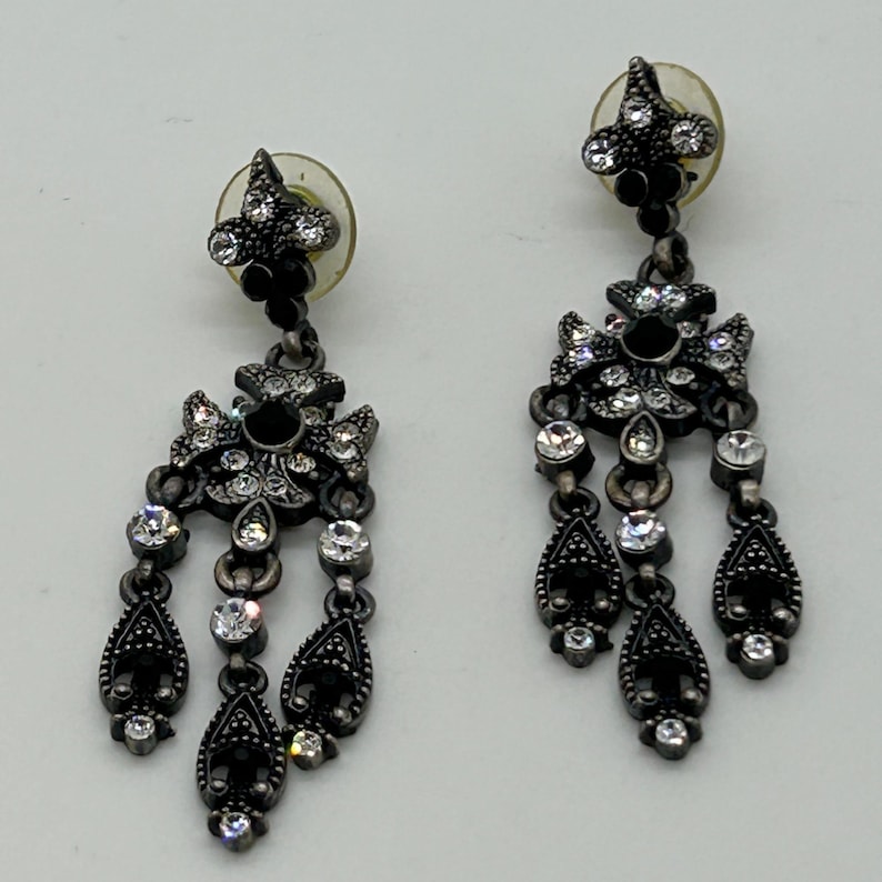 Puede incluir: Par de pendientes colgantes grises oscuros y ornamentados. Cada pendiente presenta un dise&ntilde;o floral con detalles de cristal negro y transparente. Los pendientes tienen un dise&ntilde;o en forma de l&aacute;grima.