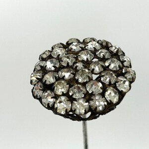 May include: A vintage hat pin with a cluster of clear, faceted rhinestones. The rhinestones are set in a dark metal filigree, forming a rounded shape. The pin has a long, slender silver-toned stem.