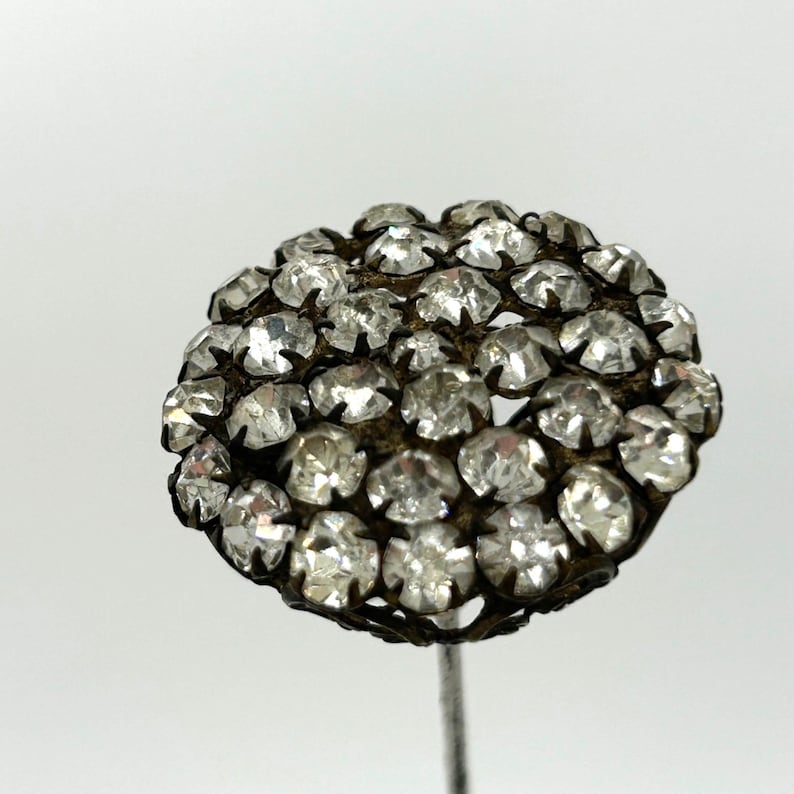 May include: A vintage hat pin with a cluster of clear, faceted rhinestones. The rhinestones are set in a dark metal filigree, forming a rounded shape. The pin has a long, slender silver-toned stem.