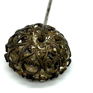 May include: Close-up of an antique silver-toned hat pin. The pin features an ornate, filigree design with a rounded, floral-like shape. The pin's metal has a tarnished appearance, suggesting age and vintage charm.