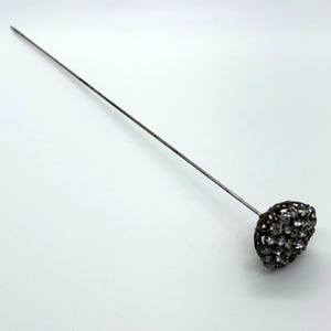May include: A silver-toned hat pin with a decorative head. The head is a cluster of small, clear crystals set in a dark metal frame. The pin is long and slender, designed to be inserted into a hat or garment.