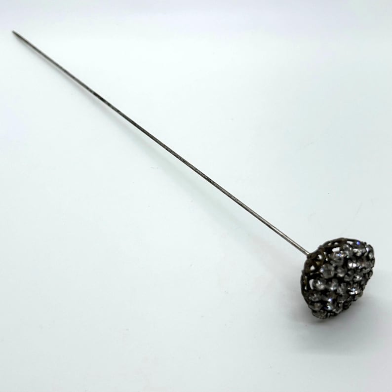 May include: A silver-toned hat pin with a decorative head. The head is a cluster of small, clear crystals set in a dark metal frame. The pin is long and slender, designed to be inserted into a hat or garment.