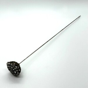 May include: A vintage hat pin with a decorative head. The head is a cluster of small, sparkling stones set in a dark metal filigree. The pin is long and slender, made of a silver-toned metal.