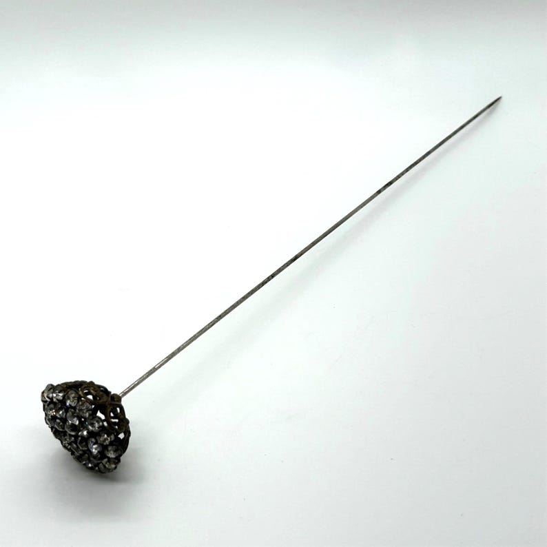 May include: A vintage hat pin with a decorative head. The head is a cluster of small, sparkling stones set in a dark metal filigree. The pin is long and slender, made of a silver-toned metal.