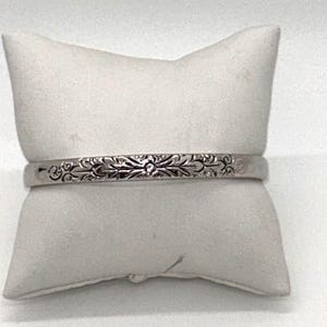 May include: A silver-toned metal bangle bracelet featuring an intricate, engraved floral pattern that spans the entire visible length of the band. The bracelet has a smooth, polished finish and a consistent width, displaying detailed scrollwork.