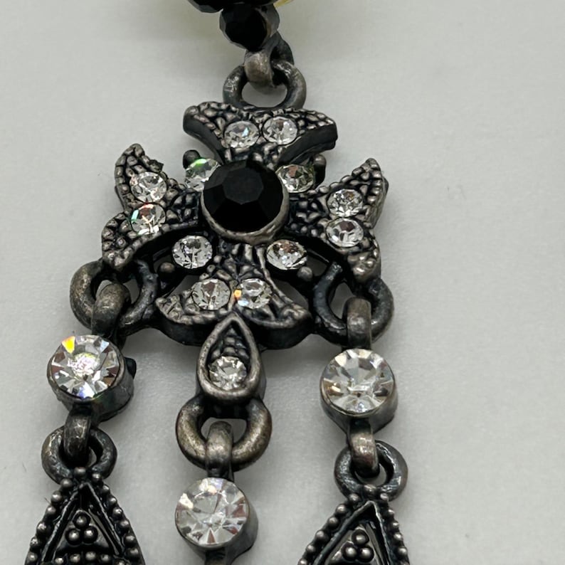 Puede incluir: Primer plano de un colgante plateado con un dise&ntilde;o floral. El colgante presenta una gran piedra negra en el centro, rodeada de cristales transparentes. Tres hilos de cristales transparentes cuelgan por debajo, lo que se suma a la apariencia ornamentada del colgante.