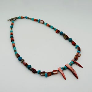 May include: A beaded necklace featuring alternating turquoise-colored round beads and reddish-brown shell fragments. The design includes small metallic spacers and three prominent, elongated, pointed shell pieces hanging from the center section.