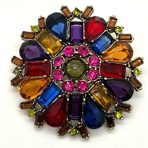 May include: A gold-toned brooch with a vibrant array of colorful gemstones. The brooch features a circular design with a central green stone surrounded by pink, red, blue, yellow, and purple gems in various shapes. Brown and green accent stones complete the design.