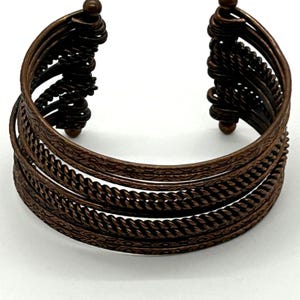 May include: Bronze-toned cuff bracelet with intricate detailing. The open-ended bracelet features multiple bands with textured and braided patterns. The ends have decorative, coiled accents and small, round finials. A stylish accessory.