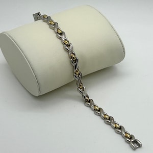 May include: A silver and gold-tone bracelet with an X and O design. The bracelet features alternating silver and gold-tone links, with small clear stones set in some of the links. The bracelet is displayed on a white surface.