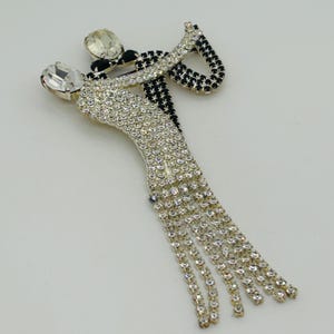 May include: A brooch shaped like two figures dancing, featuring clear and black rhinestones. One figure is covered in clear crystals with long dangling crystal fringe, while the other is accented with black stones and a large clear crystal head.
