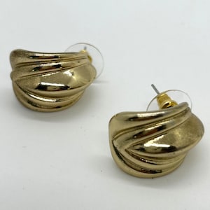 May include: A pair of gold-colored stud earrings. Each earring has a curved, ridged design, with a post and backing. The earrings are set against a white background.