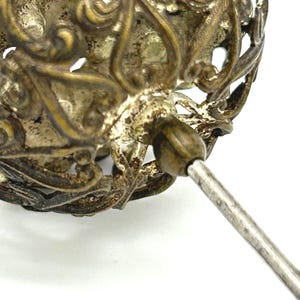 May include: Close-up of an antique silver-toned brooch. The brooch features an ornate, openwork design with a floral motif. The pin is a simple, straight design. The metal has a tarnished appearance, suggesting age and use.