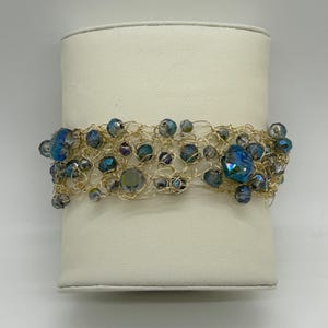 May include: A gold-toned wire bracelet adorned with iridescent blue and gray beads. The beads vary in size and shape, creating a textured and eye-catching design. The bracelet is displayed on a white surface.