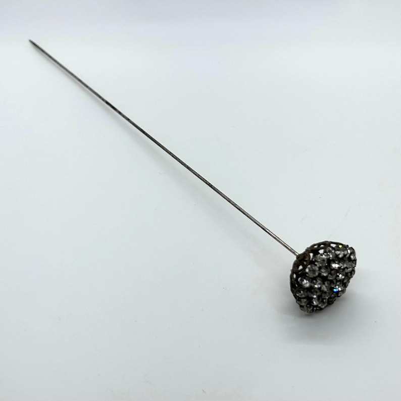 May include: A silver-toned hat pin with a cluster of small, sparkling crystals forming a rounded head. The pin is long and slender, with a simple, elegant design. The crystals catch the light, creating a dazzling effect.