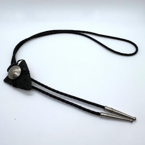 May include: A bolo tie featuring a black braided cord with silver-toned metal tips. The centerpiece is a triangular, dark, textured stone ornament adorned with a circular, engraved silver-toned metal button at its center.