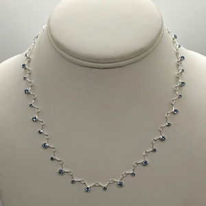 May include: A silver-toned metal chain necklace featuring a series of small, round, light blue faceted stones set at regular intervals along the links. The necklace follows a delicate, repeating pattern with each stone suspended from a short metal segment.