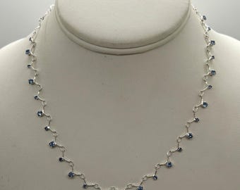 May include: A silver-toned metal chain necklace featuring a series of small, round, light blue faceted stones set at regular intervals along the links. The necklace follows a delicate, repeating pattern with each stone suspended from a short metal segment.