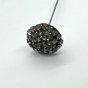 May include: A vintage hat pin with a round, dark gray head encrusted with numerous small, clear rhinestones. The pin's metal shaft extends upwards, creating a classic accessory.