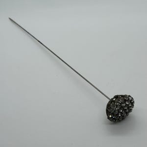 May include: A silver-toned hat pin with a rounded, textured head encrusted with small, sparkling rhinestones. The pin is long and slender, designed to secure a hat or add a decorative touch to an outfit.