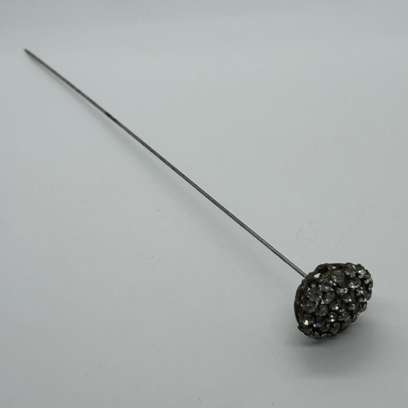 May include: A silver-toned hat pin with a rounded, textured head encrusted with small, sparkling rhinestones. The pin is long and slender, designed to secure a hat or add a decorative touch to an outfit.