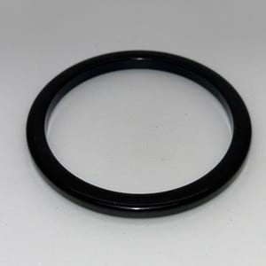May include: A glossy black bangle bracelet. The circular bracelet is a solid, smooth band. The jewelry is set against a white background. The bangle is a simple, elegant accessory.