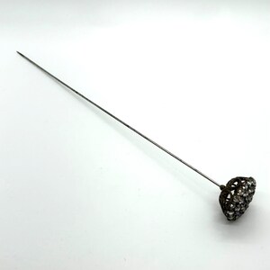 May include: A vintage hat pin with a long, slender silver-toned shaft. The decorative head is a dark metal filigree heart shape, encrusted with small, clear rhinestones. The pin is designed to secure hats or other accessories.