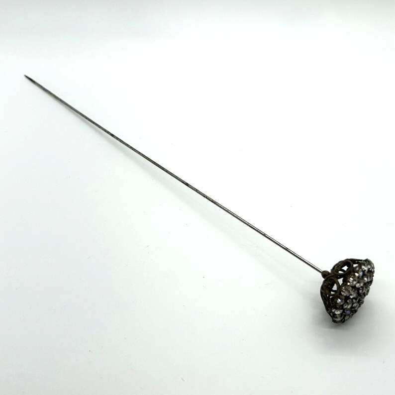 May include: A vintage hat pin with a long, slender silver-toned shaft. The decorative head is a dark metal filigree heart shape, encrusted with small, clear rhinestones. The pin is designed to secure hats or other accessories.