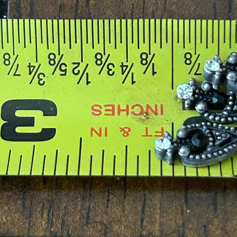 Puede incluir: Primer plano de una cinta m&eacute;trica amarilla con marcas negras y la palabra "INCHES" en naranja. La cinta mide hasta 7,6 cm. Una pieza decorativa plateada con pedrer&iacute;a est&aacute; en la esquina.