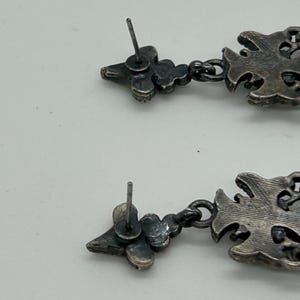 Puede incluir: Primer plano de dos pendientes colgantes plateados. Cada pendiente presenta un perno floral, conectado a una gota ornamentada. Los pendientes tienen una est&eacute;tica vintage o antigua.